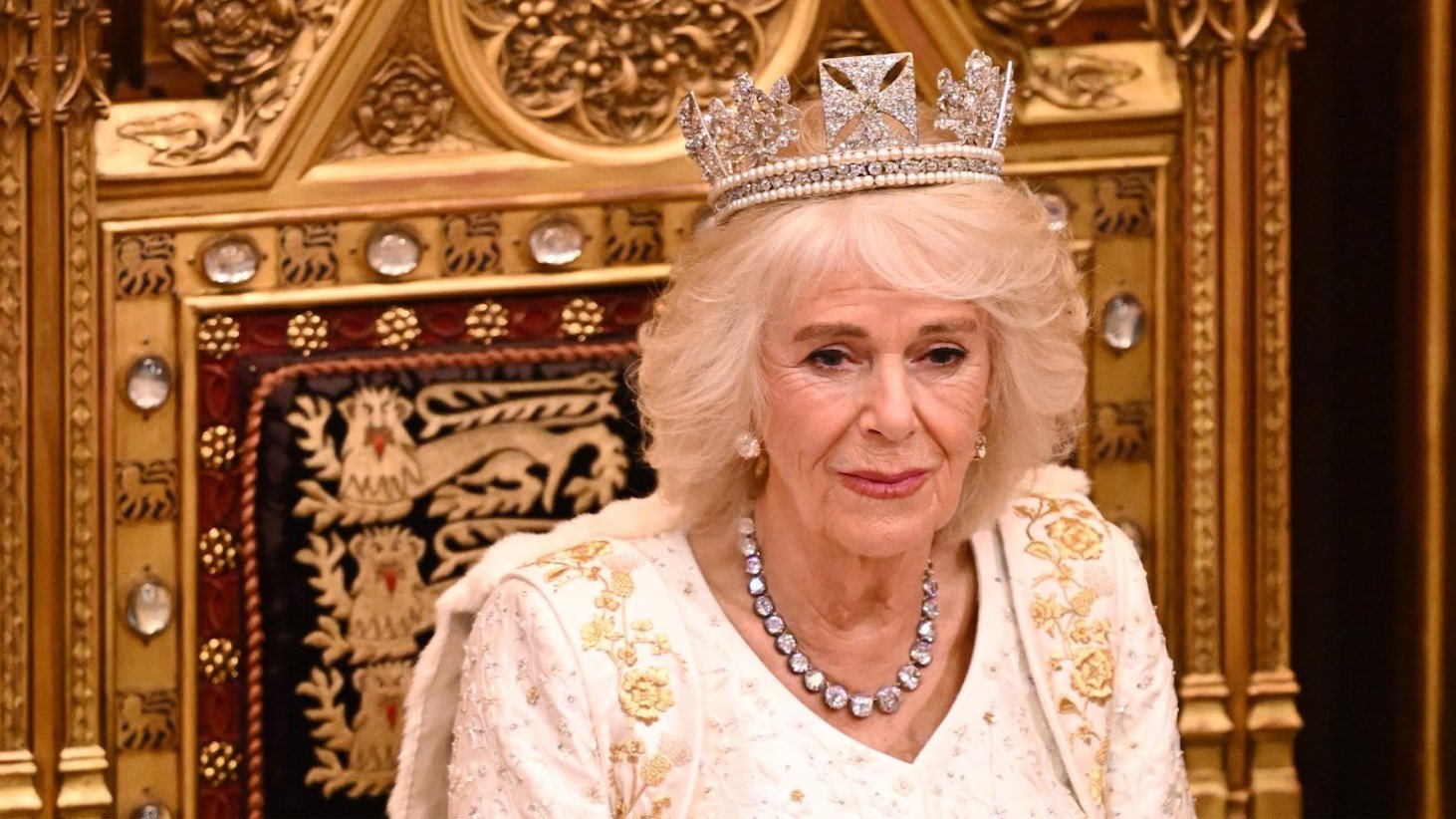 Queen Camilla is pictured wearing a ceremonial dress adorned with gold floral embroidery and a pearl necklace with sapphire accents. She dons a diamond tiara atop her white coiffed hair and is seated in front of an ornate golden backdrop with heraldic symbols