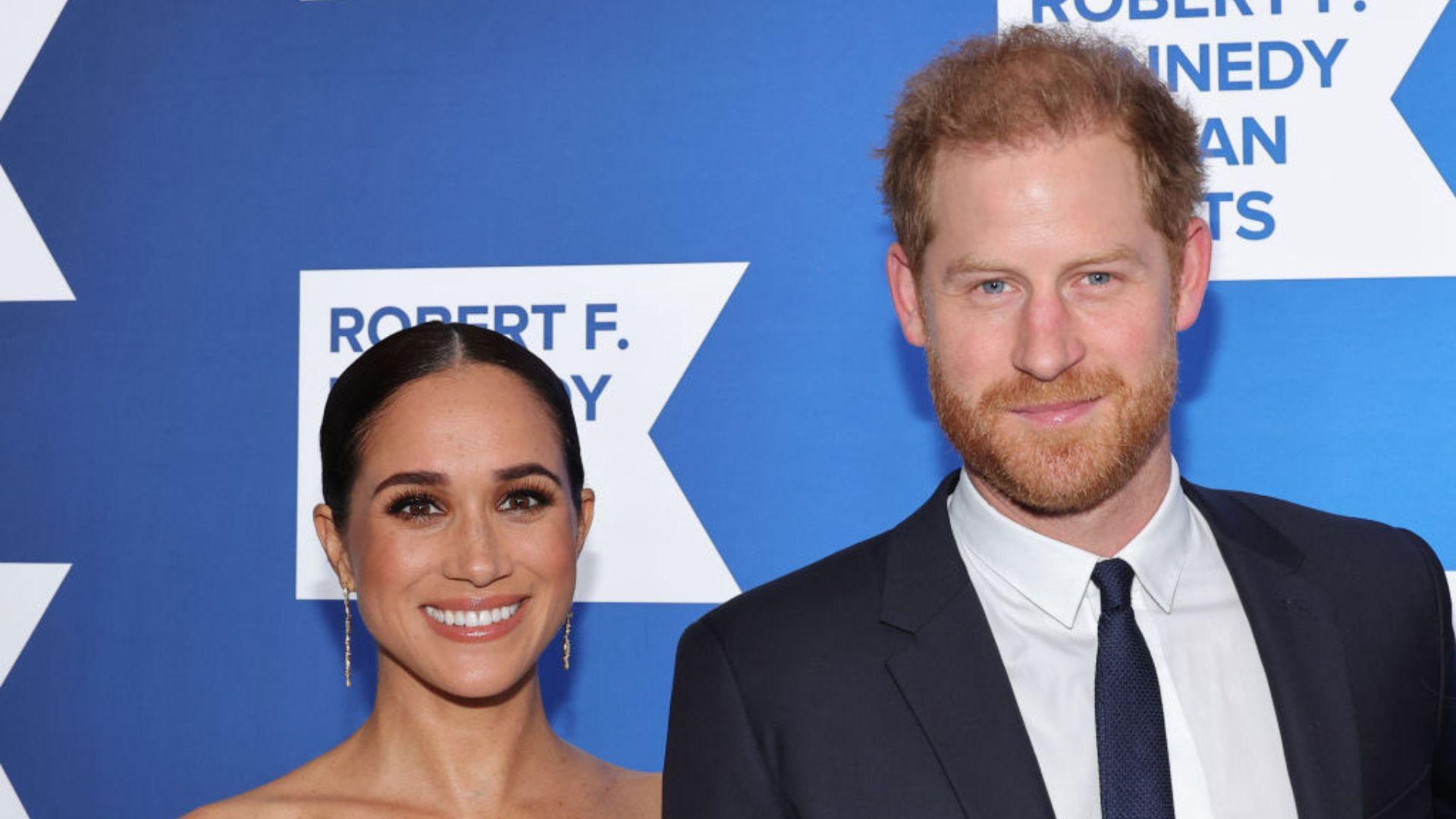Prince Harry and Meghan Markle, the Duke and Duchess of Sussex, are smiling and posing together for a photo at a formal event. Meghan is wearing a sleeveless dress with her hair neatly tied back, complemented by long earrings, while Prince Harry is in a dark suit with a light shirt and a dark blue tie